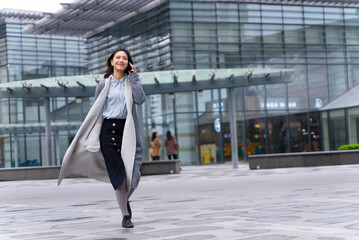 Young beautiful girl walking outside office building using mobile phone and happily calling customers