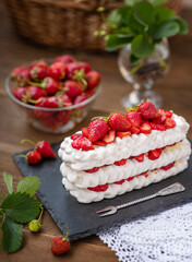 Pavlova dessert with strawberries and violet flowers as decor on a wooden background next to a plate full of strawberries