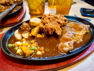 Close up thin grilled sirloin beef steak on a hot plate with fried chicken, potato, vegetables and black pepper sauce at indonesian restaurant.