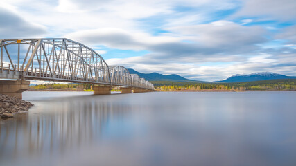 Obraz premium Large man made structure steel bridge spanning across Nisutlin Bay in township of Teslin flowing to the Yukon River in northern Canada during spring summer time with cloudy mountains background.