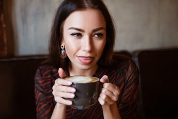 Portrait of a young woman of Caucasian appearance in a cafe with a cup of coffee. Beautiful brunette with brown eyes. Enjoying morning coffee in a cozy cafeteria