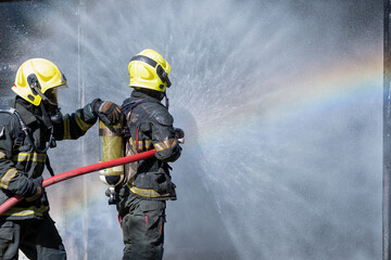 Fototapeta premium Two fireman or firefighters stand and help each other to spray of water with curtain shape and rainbow reflex occur on water aerosol in front of container containing fire inside.