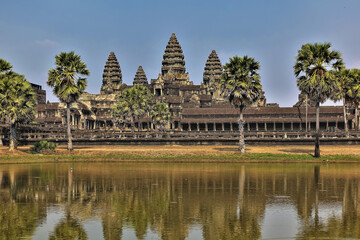 Fototapeta na wymiar Ancient temple of Cambodia Angkor Wat. High carved towers against the background of the sky. Colonnades and galleries are dilapidated. Palm trees on the shore of the lake. Reflection in water.