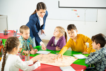 Group of school kids with teacher sitting together around desk in classroom, playing educational...
