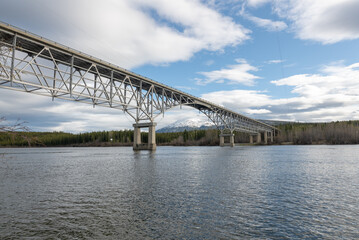 Johnsons Crossing, Teslin River steel Bridge on the Alaska Highway during spring summer time with cloudy, blue sky day and magnificent, huge structure over the flowing water below. 