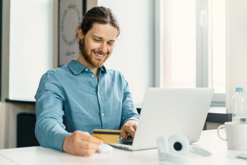 Fototapeta premium Happy young man sitting at desk in office with credit card and shopping online on laptop, joyful male buyer making payment on computer online, using internet banking service system