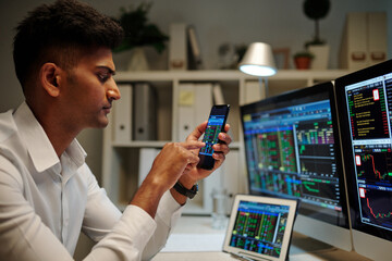 Serious broker checking stock soar via application on smartphone when working in dark office
