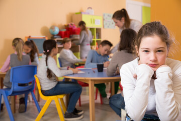 Portrait of sad ordinary girl and children drawing in classroom