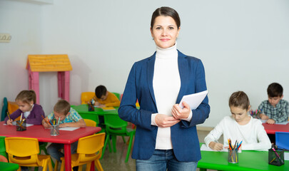 Portrait of friendly female teacher standing in classroom on background of elementary school students
