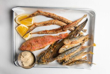 Fried fish with sauce and lemon served on a metal tray over white background.