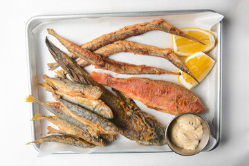 Fried fish with sauce and lemon served on a metal tray over white background.