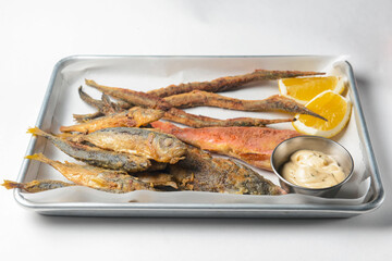 Fried fish with sauce and lemon served on a metal tray over white background.