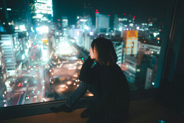 窓際で東京の夜景を眺めながら黄昏れている女性