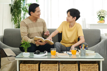 Father and son dicussing difficult topic and examples in students book when sitting on sofa at home
