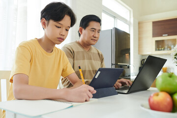 Concentrated teenage boy doing homework at kitchen table when sitting next to his father working on laptop
