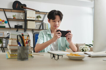 Cheerful teenage boy playing game on smartphone instead of doing homework at home