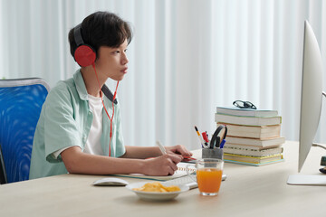 Concentrated serious schoolboy in headphones watching educational video on computer and writing in notepad