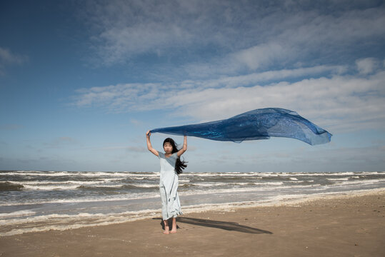 Asian girl in blue dress on the beach near ocean holding thin fabric in the wind