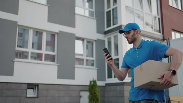 Caucasian young pretty man, delivery worker in blue cap walking the street and carrying carton box while using smartphone, looking for route. Male courier with parcel tapping and texting on phone.