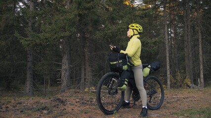 The woman travel on mixed terrain cycle touring with bike bikepacking outdoor. The traveler journey with bicycle bags. Sportswear in green black colors. Magic forest park. Make a selfie smartphone.