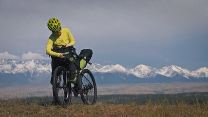 The woman travel on mixed terrain cycle touring with bikepacking. The traveler journey with bicycle bags. Sport tourism bikepacking, bike, sportswear in green black colors. Mountain snow capped. - obrazy, fototapety, plakaty