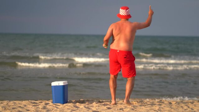 Chubby man in front of the sea with a camping fridge and starts dancing