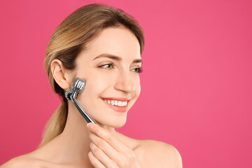 Young woman using metal face roller on pink background