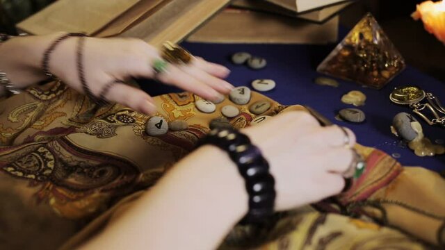 Astrology and esotericism. A female fortune teller makes a layout with Runestones, using a book. Hands in rings and bracelets close-up.