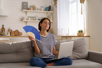 Tired millennial woman suffers from stuffiness and an inoperative air conditioner, waving blue fan...