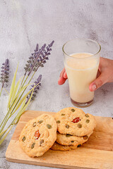 A hand is picking up a glass of milk. cereals cookies are stacked on cutting board, cookies topped with almonds, raisins, sunflower seeds, pumpkin seeds, whole grains.