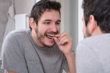 Young latin man putting orthodontic silicone trainer or invisible braces aligner in front of the mirror. Mobile orthodontic appliance for dental correction.