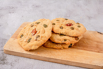Close up of cereals cookies are stacked on cutting board, cookies topped with almonds, raisins, sunflower seeds, pumpkin seeds, whole grains.
