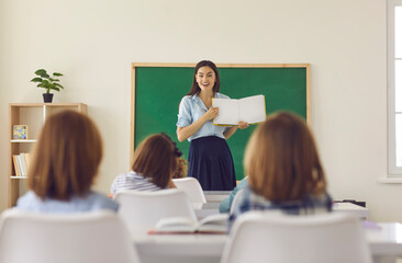 Teacher conducts a lesson at school. Young friendly Caucasian female teacher stands in the classroom by the blackboard with a book in her hands and tells something interesting to the little students.