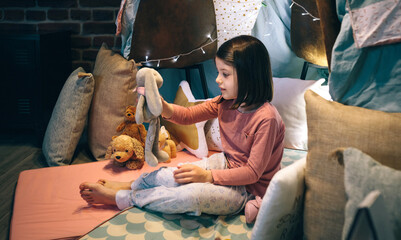 Adorable girl in pajamas playing alone with teddies in a play shelter made of bed sheets and chairs