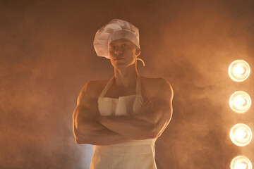 Portrait of muscular chef wearing white apron and chef hat, posing with folded arms on smoky...