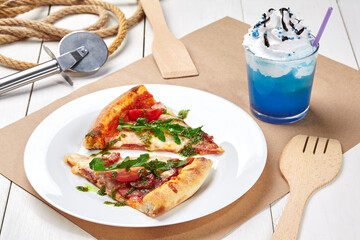 Two slices of pizza with sausages, tomatoes, greens served with blue lemonade