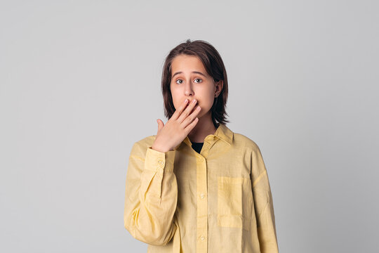 Portrait of beautiful teen girl keeping with hand over mouth, saying oops wearing casual yellow shirt, gray background
