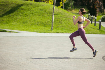 Fototapeta premium Young female runner, athlete is jogging in the city street in spring sunshine. Beautiful caucasian woman training, listening to music. Concept of sport, healthy lifestyle, movement, activity.