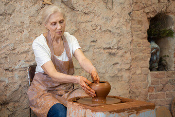 Fototapeta premium Elderly female potter working on pottery wheel while sitting in her workshop