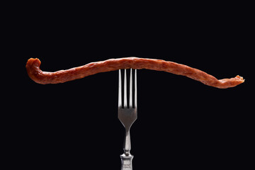 Closeup of long thing sausage on the steel fork against black background