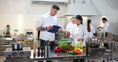 Asian pretty professional female cook stands at restaurant kitchen choosing and ordering food products talking to Caucasian man co-worker. People working on background. Culinary, job concept