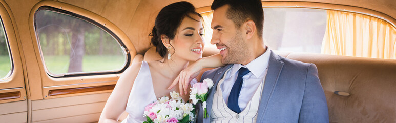 Naklejka premium Cheerful bride with bouquet looking at groom in vintage auto, banner
