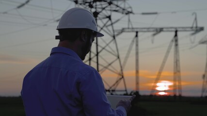 Power engineer in white helmet checks power line using computer on tablet, remote control of power system. High voltage electrical lines at sunset. Distribution and supply of electricity. Energy
