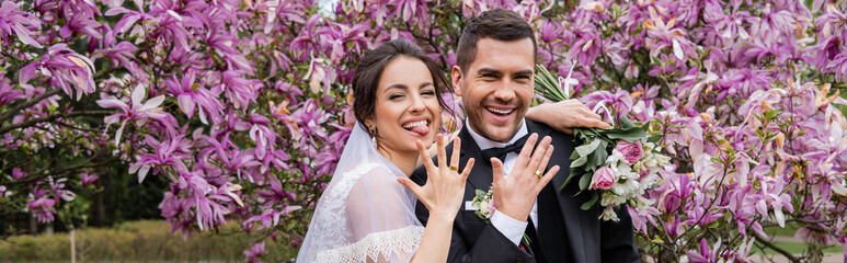 Cheerful bride and groom showing rings and looking at camera near magnolia trees, banner