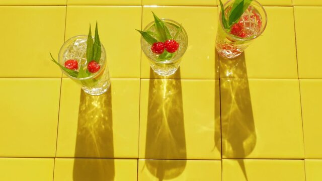Refreshing summer drinks in crystal glasses with green leaves raspberry yellow tile background shadows. Sweet cold natural cocktail hot summer day. Soda water fruits berries mint tonic. Party vacation