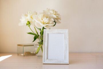 Portrait white picture frame mockup on wooden table. Modern glass vase with peony. White wall background. Scandinavian interior. Vertical.
