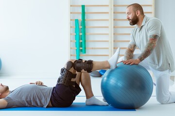 Young professional physiotherapist exercising with an injured patient using a blue ball