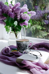 Lilac bouquet, an open book and a cup of tea on a window sill.