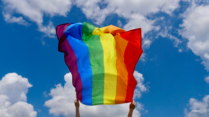 Female hand holding a LGBT gender identity flag on a background of blue cloudy sky on a sunny windy summer day during the celebration of the month of dignity at the parade