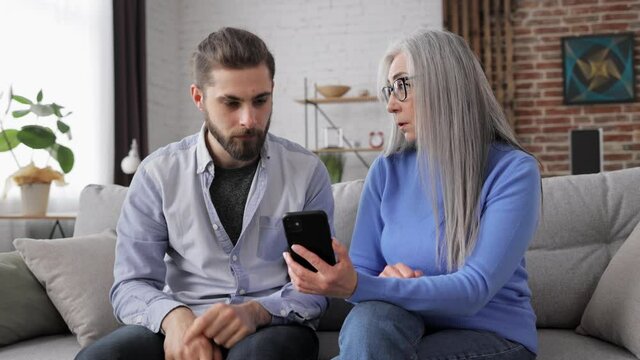 Adult son teaching his senior mother to use smartphone mobile apps. Mom does not understand expalanations, son feeling nervously. Family and generations conflicts, misunderstanding.